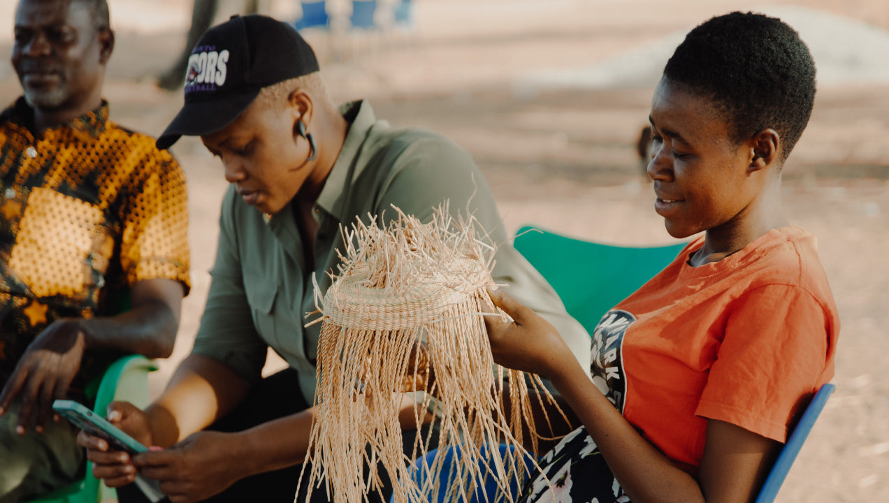 a woman sitting outdoor and weaves a Frafra basket, next to other woman with phone